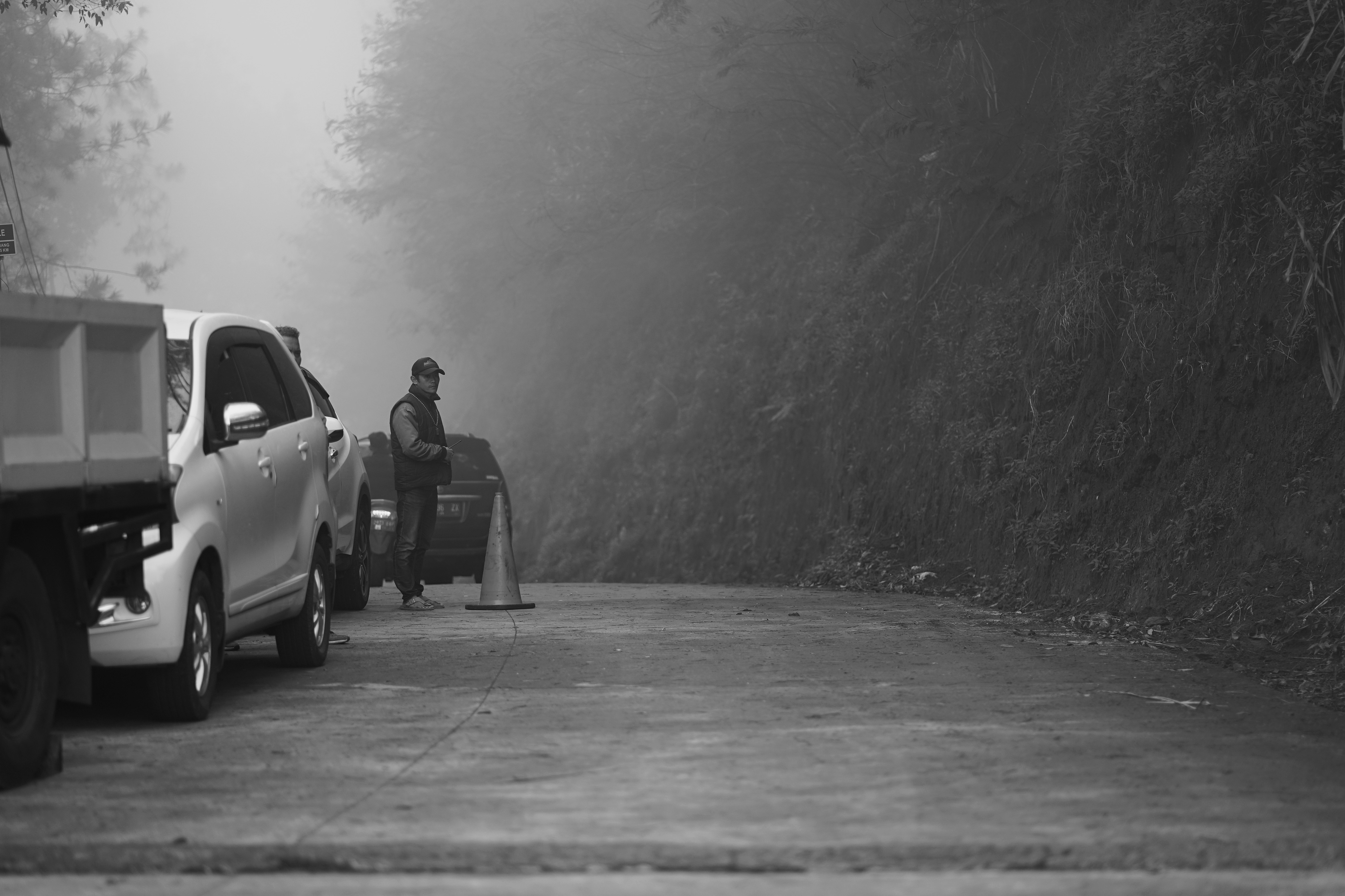 grayscale photo of man in black jacket and pants standing beside car